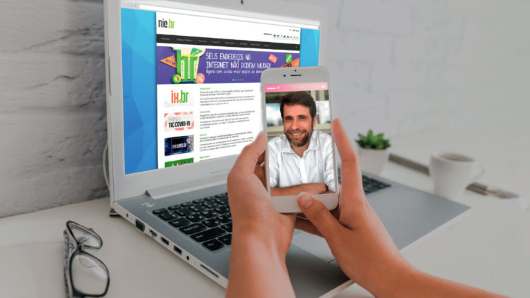 Acessibilidade na Web. Imagem de duas mãos segurando um celular com a foto do entrevistado Reinaldo Ferraz. Atrás há um notebook acessando o site do Núcleo de Informação e Coordenação do Ponto BR - NIC.br.