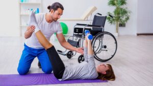 Fisioterapeuta homem atendendo mulher cadeirante, que está deitada realizando exercício, pelo Dia Nacional do Físio e do T.O. 2020