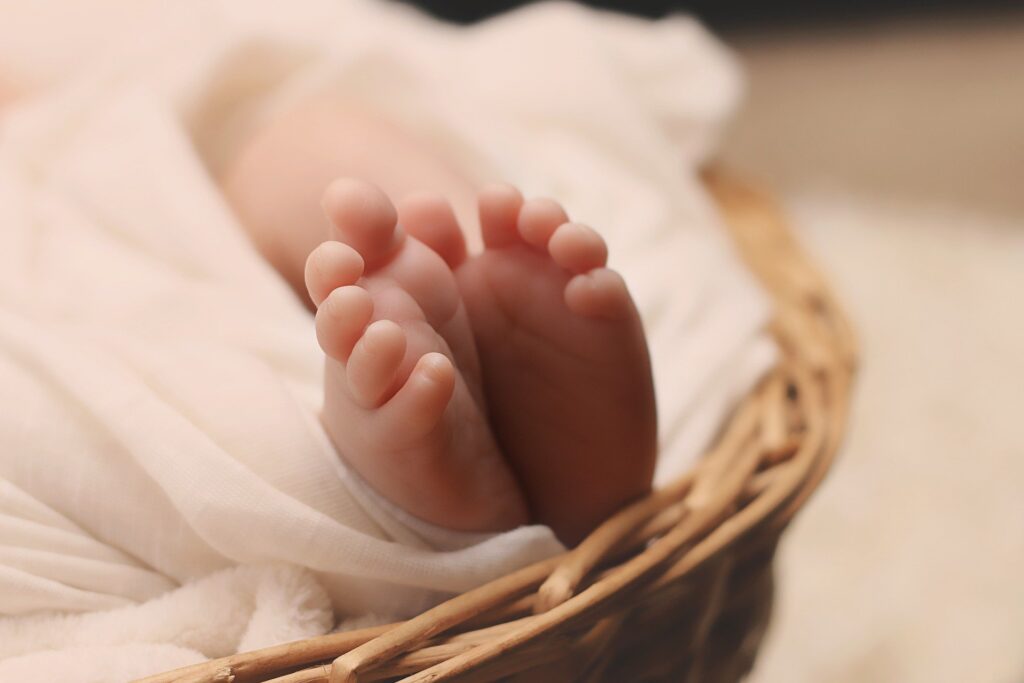 Dois pezinhos de um bebê, dentro de um cesto de palha. Tem a nele branca e está envolto a um cobertor branco.