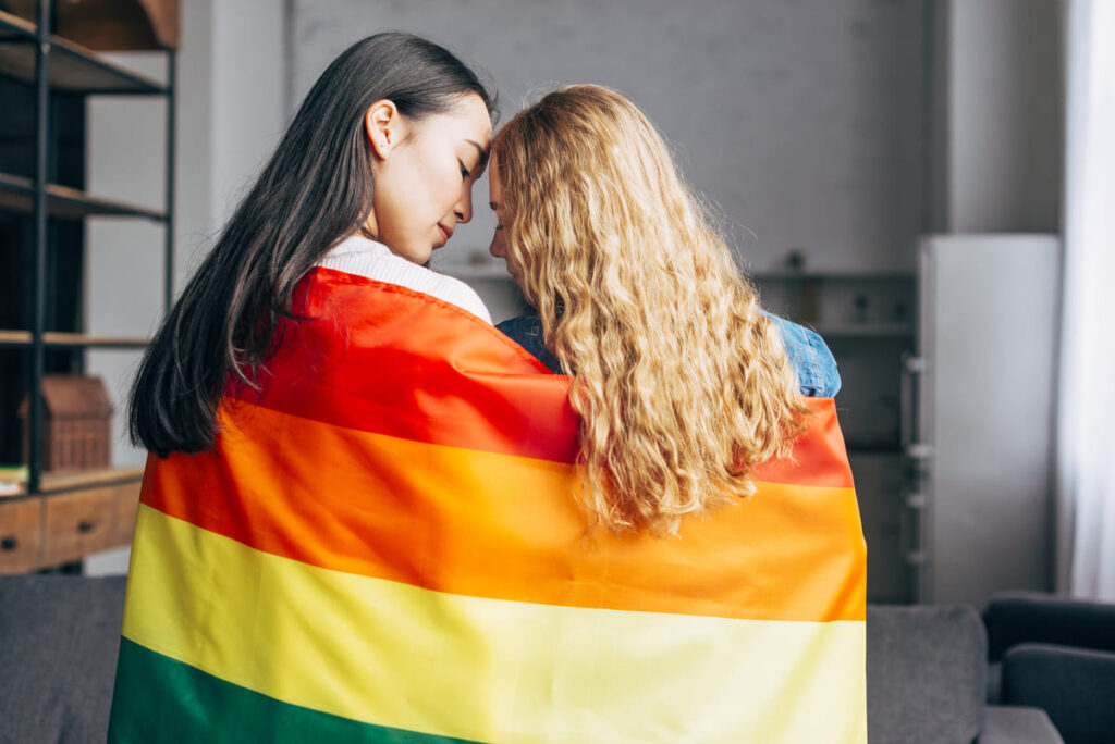 Duas mulheres juntas com a bandeira LGBTQIA+, descritas na legenda.