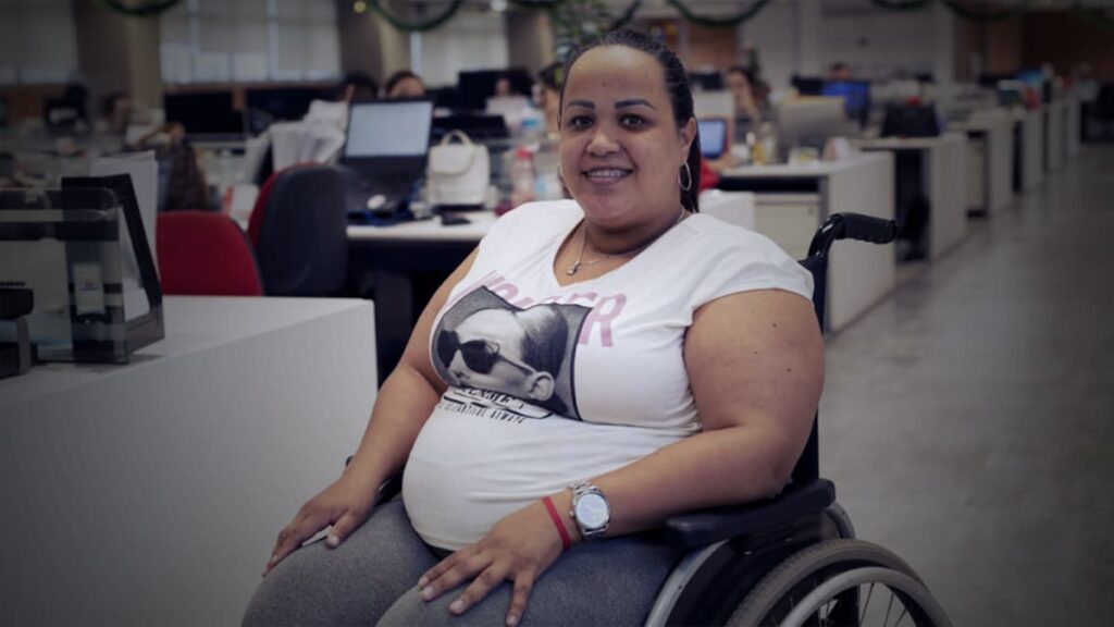 Foto de Gabriela Peña, mulher em cadeira de rodas, em ambiente corporativo. Descrição detalhada na legenda.