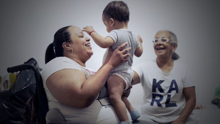 Foto de mulher em cadeira de rodas, carregando o bebê, ao lado da avó, com descrição na legenda do texto sobre "cadeirante refugiada da Venezuela".