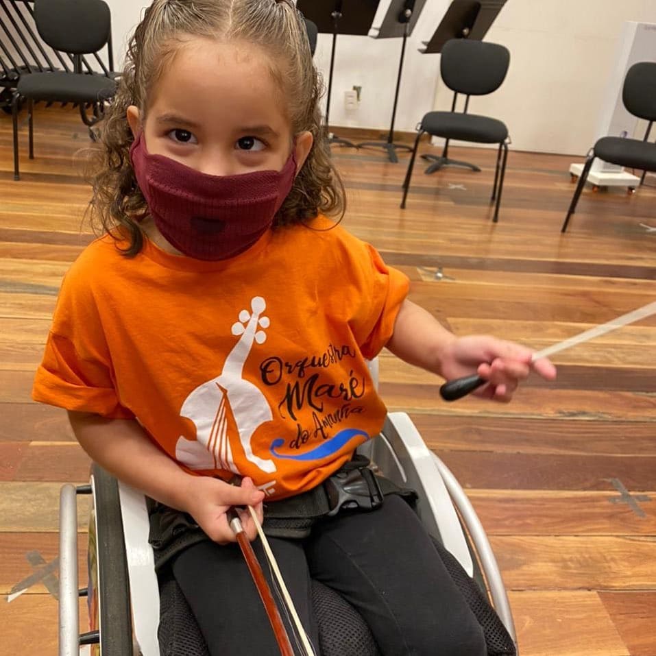 Projeto mostra a importância da música na vida de crianças com deficiência 4 Criança branca de cabelos cacheados, sentada na cadeira de rodas, durante intervalo de ensaio da orquestra.
