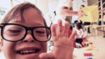 Criança usando óculos de grau, interage com fotógrafo, para ilustrar 10 sinais de alerta para detectar o TEA.