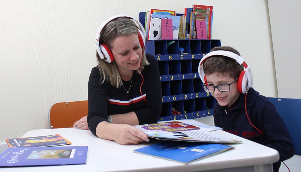 Criança com baixa visão fazendo leitura de audiobook com auxilio de mulher adulta.