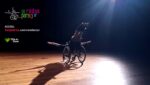 Foto de apresentação de espetáculo com artista em cadeira de rodas e sobreposição da logo De Rodas para o Ar, do Dia de Doar, e do link da campanha de arrecadação, detalhados na legenda abaixo.