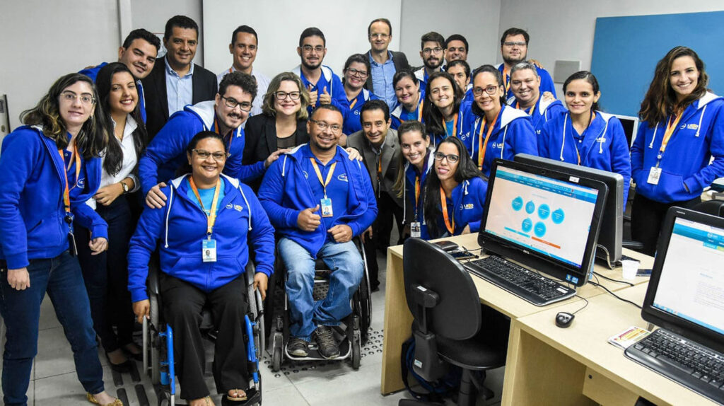 Foto colorida, em ambiente interno, com diversas pessoas, entre homens e mulheres, com e sem deficiência, da equipe do Centro de Pesquisa, Desenvolvimento e Inovação Dell (LEAD).