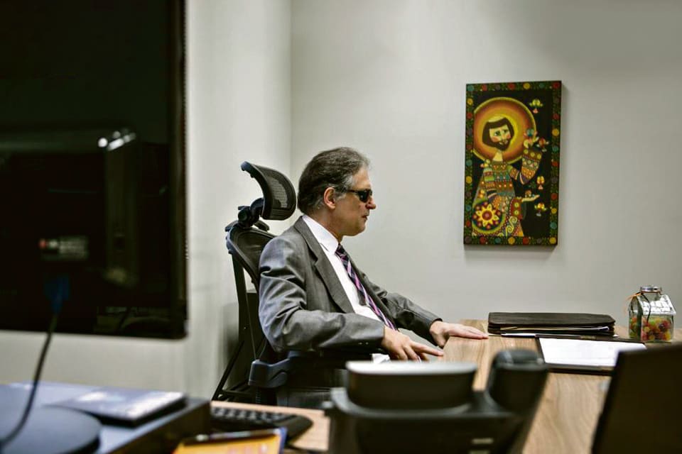 Desembargador cego, Ricardo Tadeu, homem branco de cabelos grisalhos, terno e gravata, e óculos escuros. Está no escritório.