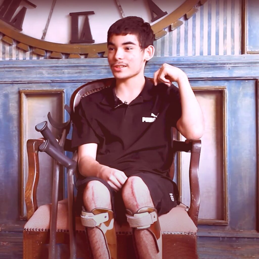 Foto do Pedro, adolescente com pele branca e cabelos pretos curtos. Está sentado, usa órteses em membros inferiores e tem duas muletas apoiadas ao seu lado.