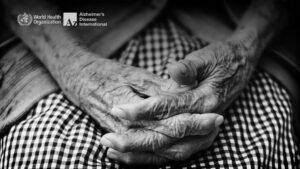 Fotografia em preto e branco, mostra em detalhes as mãos de uma pessoa idosa, com os dedos entrelaçados, acima das pernas, ilustrando a Comunidade Amiga da Pessoa com Demência.