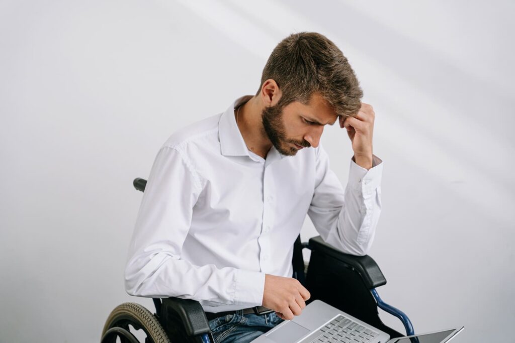 Homem branco, com a cabeça baixa, sentado em cadeira de rodas com notebook no colo.