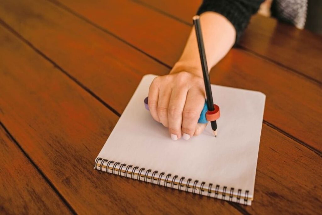 Uma mão feminina segurando um engrossador horizontal, que está acoplado a um lápis e sobre um caderno.