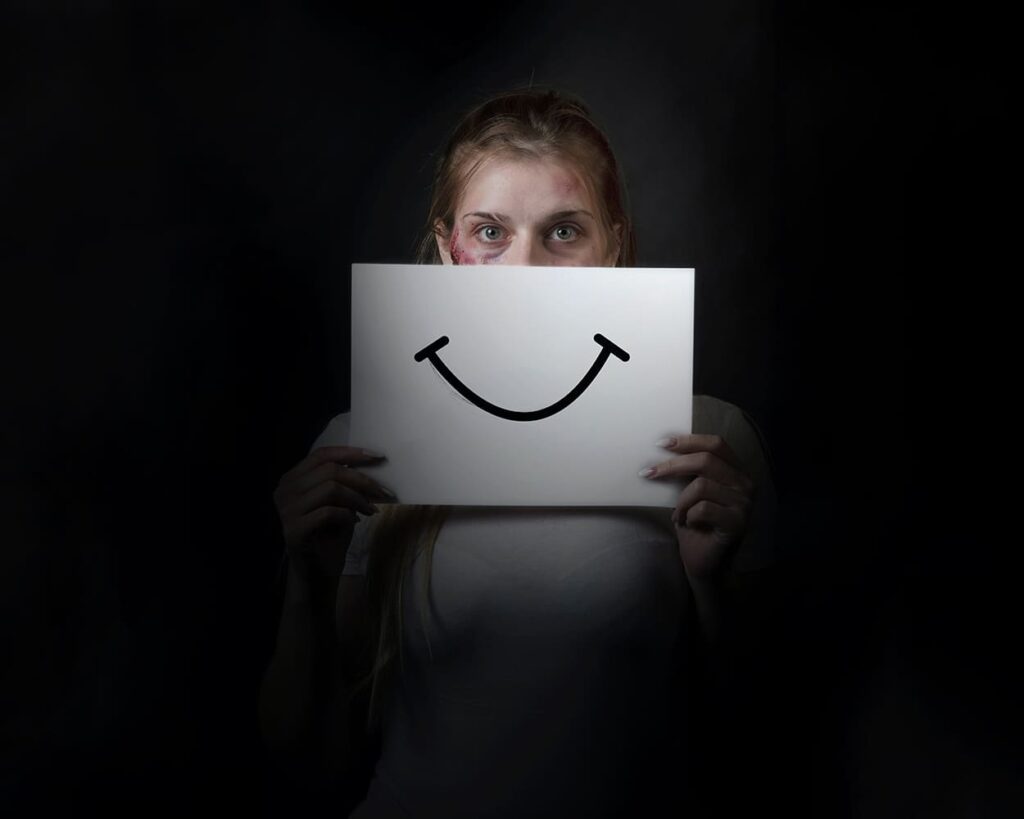 Foto em ambiente escuro, de pessoa com o rosto machucado, segurando um papel com o desenho de sorriso.