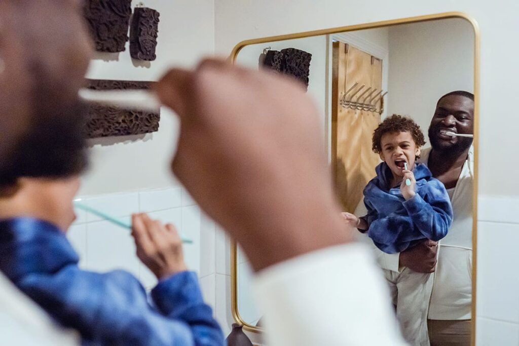 Homem negro carrega o filho de frente para o espelho, ambos escovando os dentes.