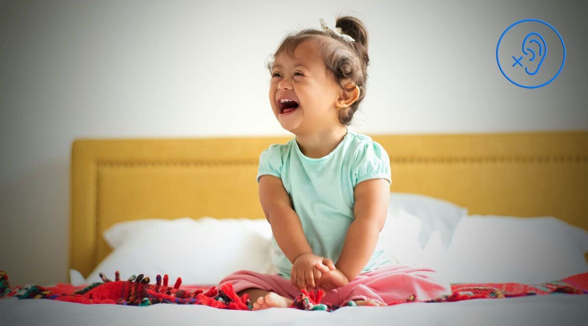 Criança parda com perda auditiva na trissomia 21 – síndrome de Down, sentada na cama e sorrindo.