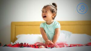 Criança parda com perda auditiva na trissomia 21 – síndrome de Down, sentada na cama e sorrindo.