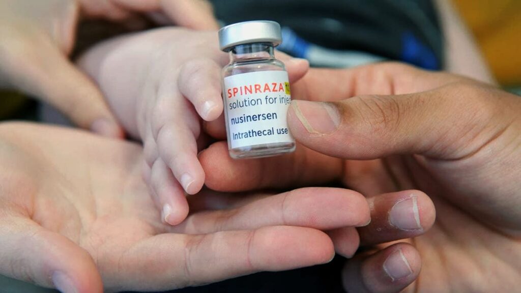 Foto com mãos de adultos e de uma criança segurando frasco do medicamento Spinraza.