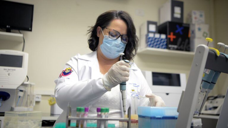 A imunologista pediátrica Carolina Prando, mulher branca com cabelos pretos curtos, manejando instrumentos no laboratório. Ilustra a manchete APS1: Irmãos descobrem doença rara após hospitalização por COVID-19.