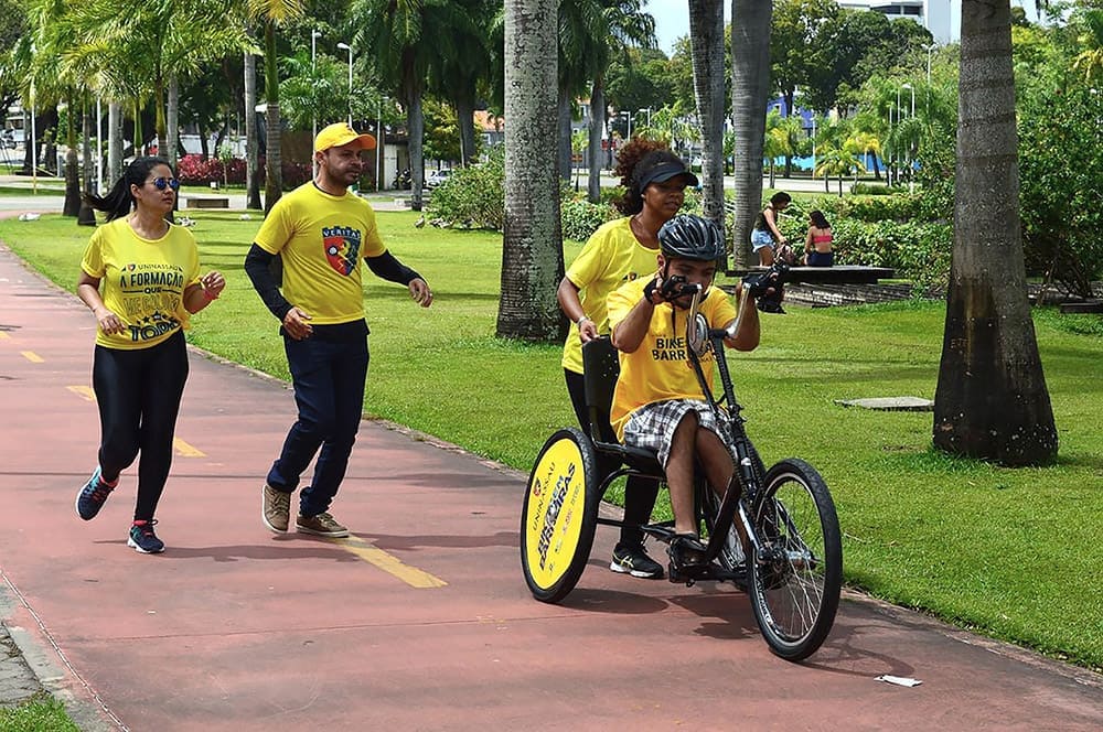 Quatro pessoas participam do projeto Bike Sem Barreiras. Tres estão correndo a pé e a quarta pessoa, que tem deficiência, pedala uma handbike com as mãos.