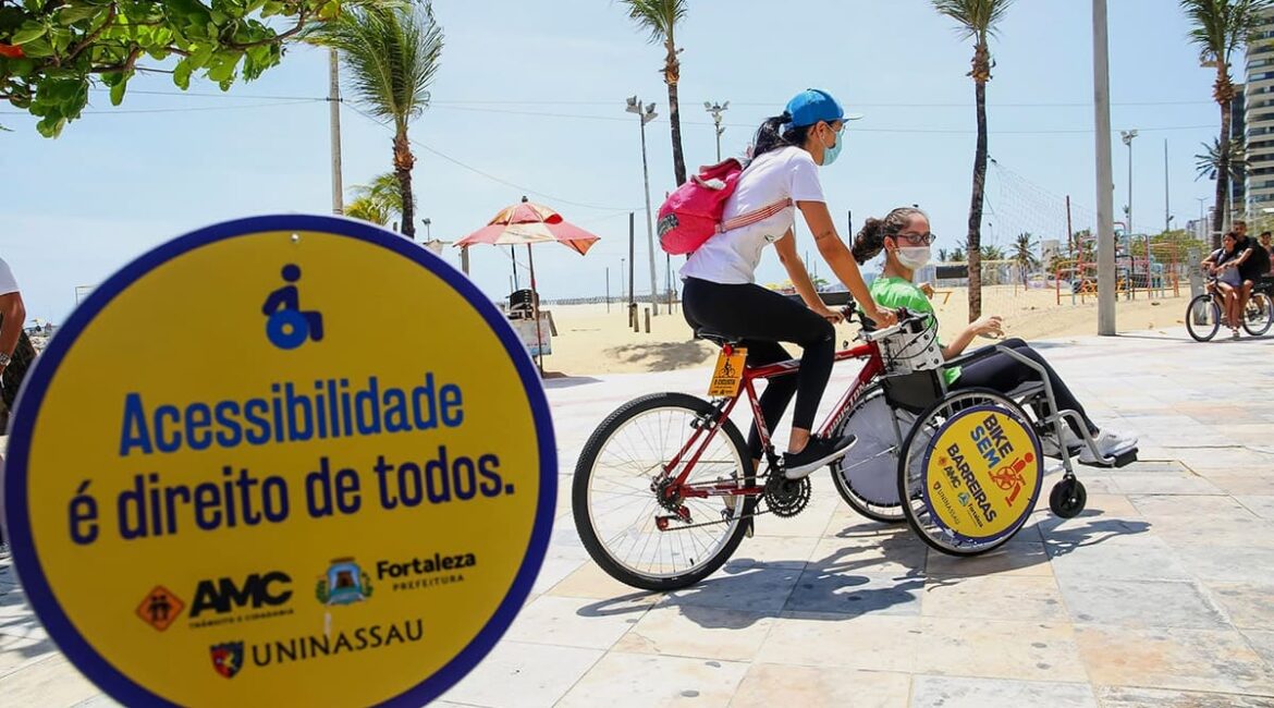 Foto do Bike Sem Barreiras, projeto de bike adaptada para PcDs, com duas pessoas: Uma em cima da bicicleta e outra na cadeira de rodas, que serve como roda frontal da bike.