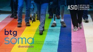 Pessoas atravessando a rua na faixa de pedestres, com as cores do arco-íris e sobreposição dos nomes BTG Soma Diversidade e ASID – Ação Social para Igualdade das Diferenças.
