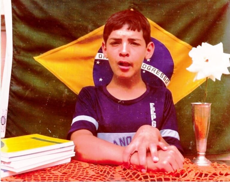 Emilio Figueira em 1984, no colégio. Tem pele clara e cabelos escuros e curtos. Está sentado e com as mãos sobre a mesa. Ao fundo está a bandeira nacional.