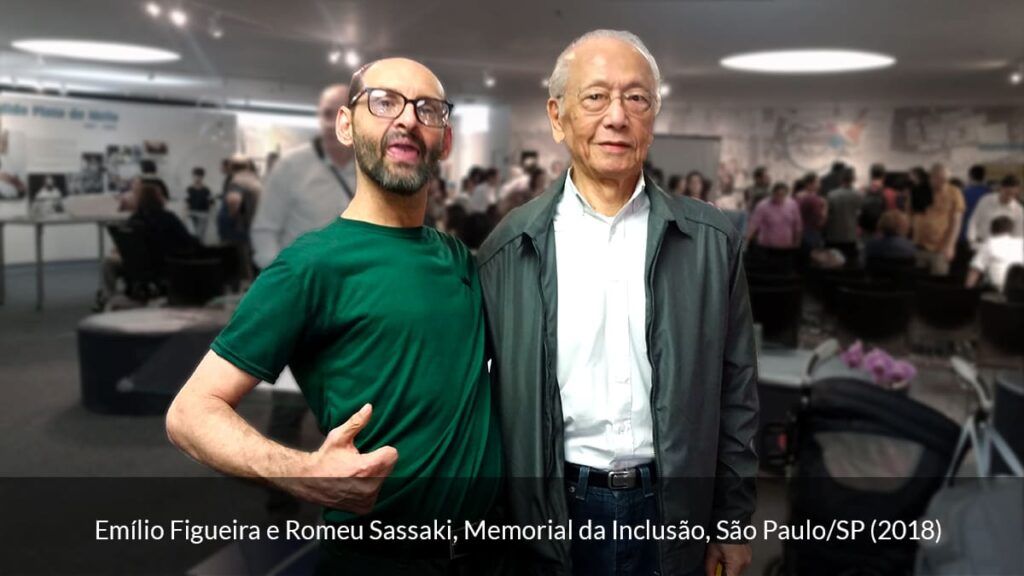 Emílio Figueira, homem calvo com óculos e camiseta verde. Está em pé, ao lado de Romeu Sassaki. Homem branco de cabelos grisalhos, óculos, camisa branca e casaco, em 2018.