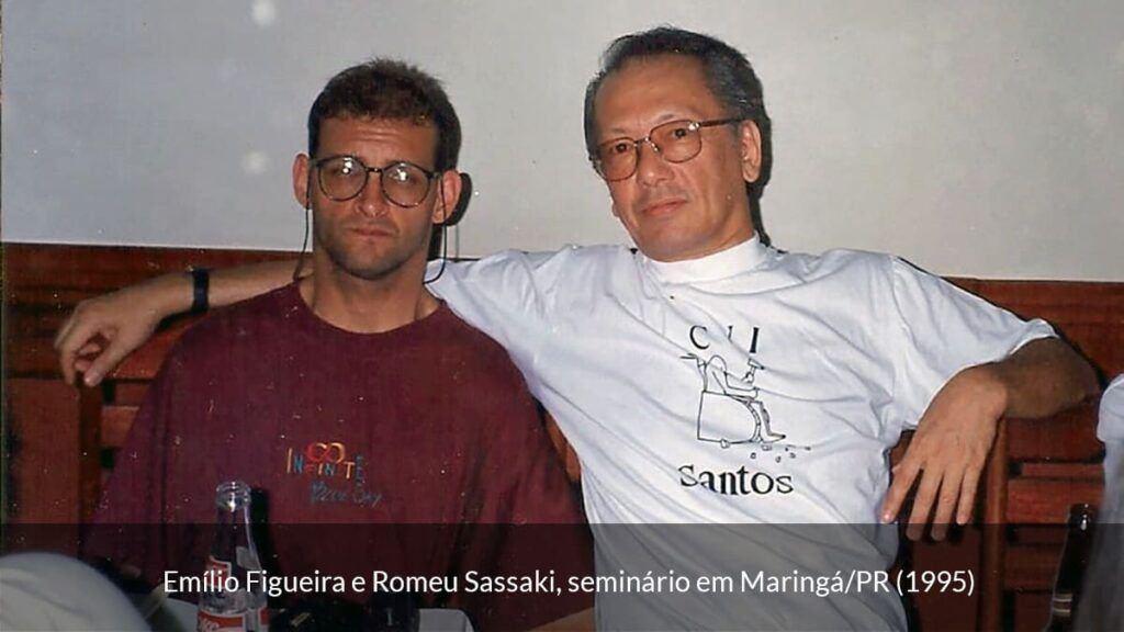 Emílio Figueira, homem branco com óculos e camiseta bordô. Sentado ao lado de Romeu Sassaki. Homem branco de óculos e camiseta branca, em 1995.