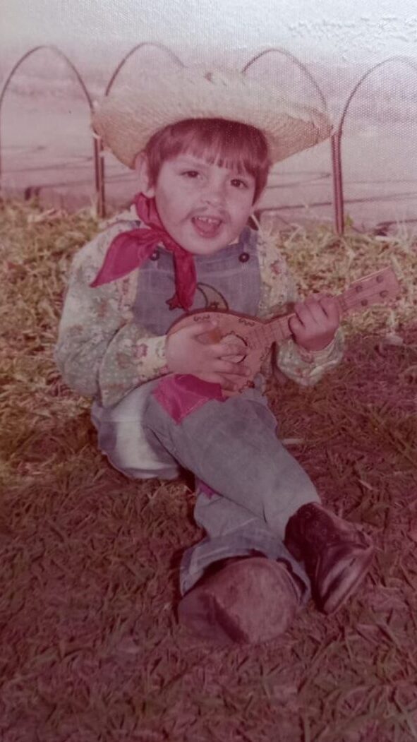 Emilio Figueira criança, sentado na grama. Usa vestes de São João e toca um pequeno violão.