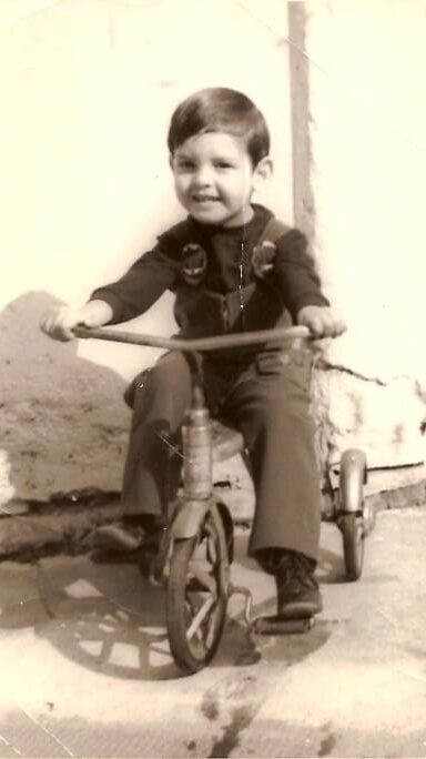 Emilio Figueira criança pedalando um triciclo. Sorri e usa macacão.