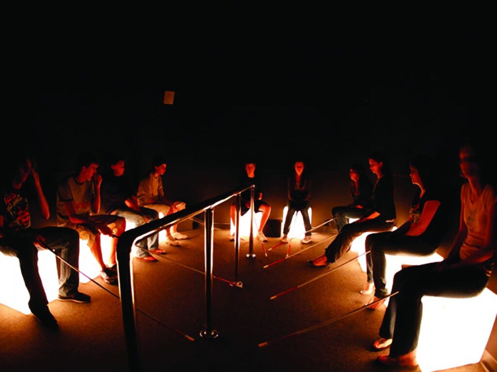 Foto de grupo de pessoas em ambiente escuro da exposição. Estão sentadas segurando bengalas utilizadas por pessoas cegas