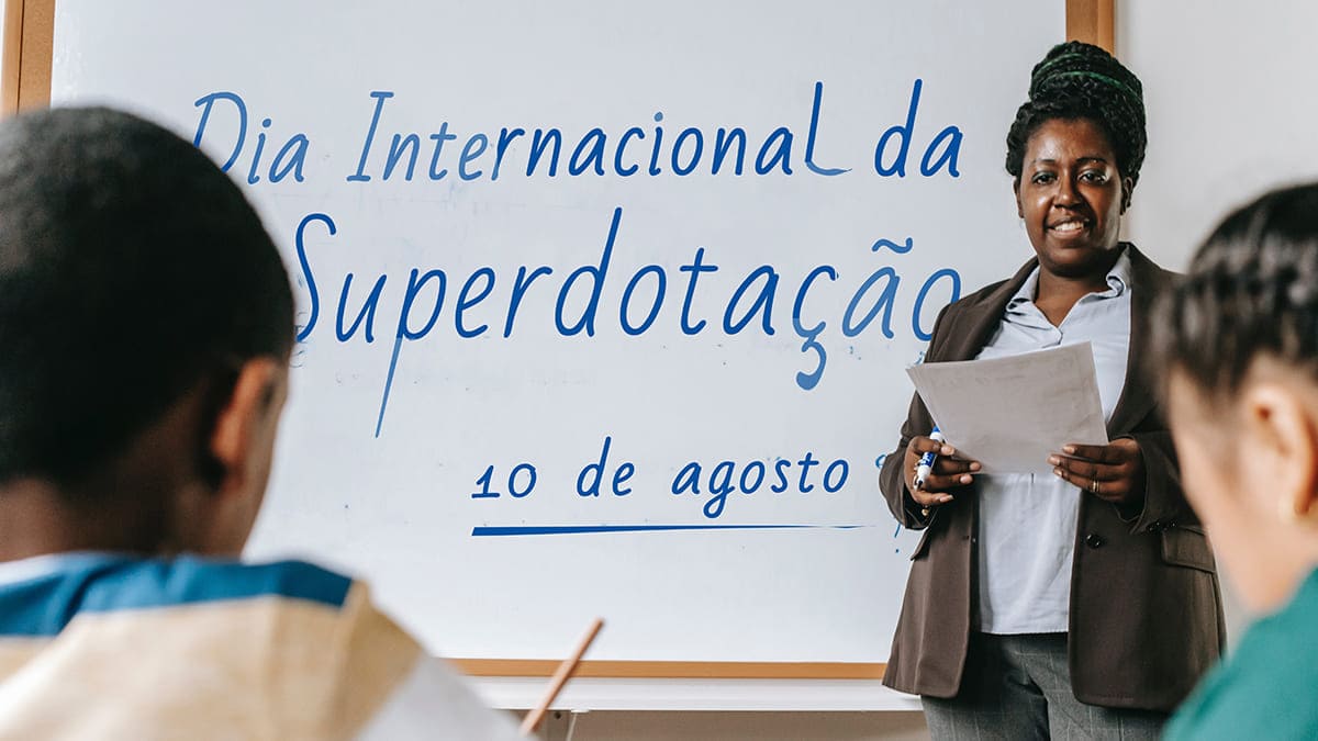 Superdotação no Brasil: Foto em sala de aula com professora negra e texto na lousa: Dia Internacional da Superdotação, 10 de agosto.