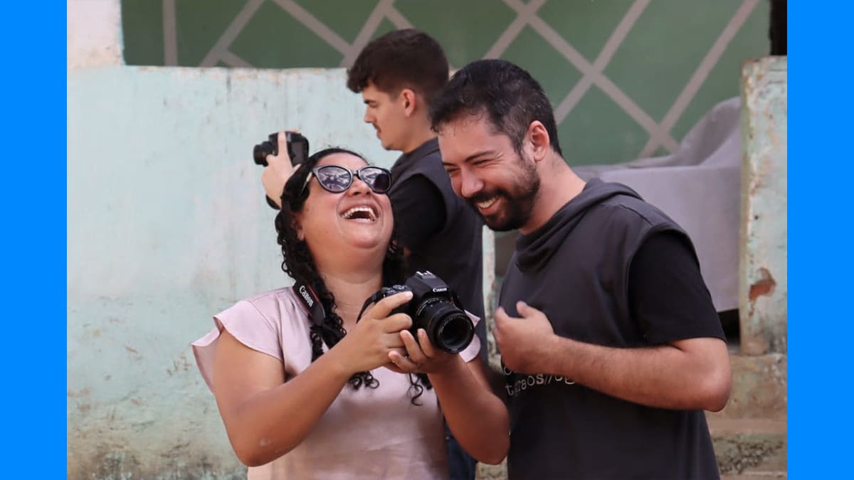 Foto colorida com Julio e Sayonara. Ela segura máquina fotográfica e os dois estão rindo. Ilustra a pauta dos 12 fotógrafos cegos no Espírito Santo.