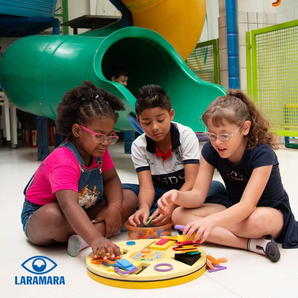 Foto em ambiente interno com três crianças brincando e sobreposição do logo da Laramara, no canto esquerdo inferior: Brincadeiras para crianças com deficiência visual.