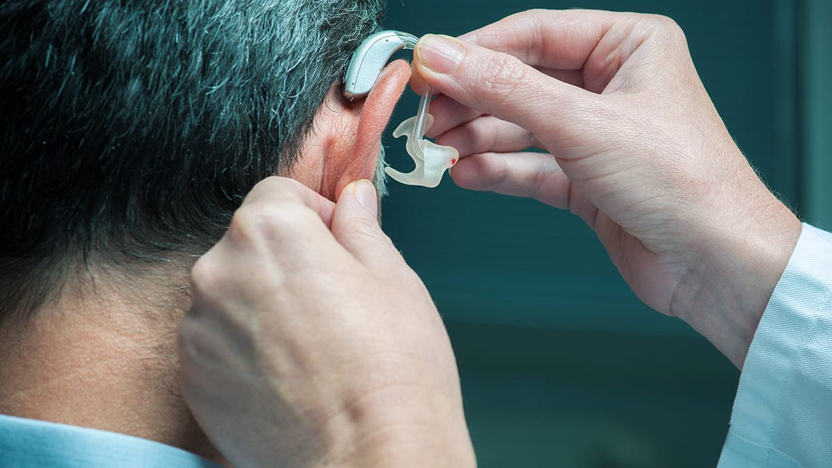 PL 2225 Deficiência auditiva unilateral, pessoa branca colocando o aparelho de audição na orelha direita de outra pessoa, que está de costas.