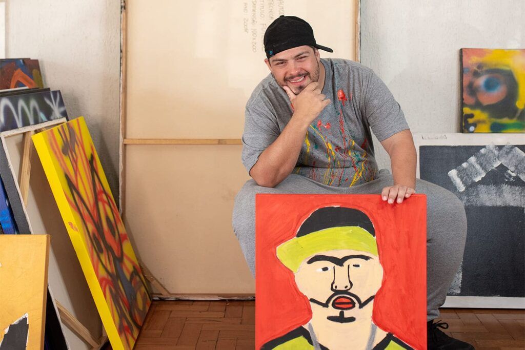 Artista plástico com T21, Lucio Piantino está sentado com o queixo apoiado na mão direita e sorrindo. À sua frente segura o autorretrato. Dos lados há outras várias telas.