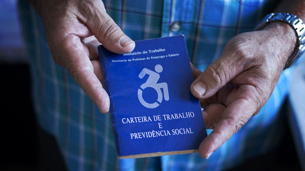 Mãos seguram carteira de trabalho com símbolo de cadeira de rodas sobre o brasão do Brasil na capa. Auxílio de até 1 salário mínimo a desempregados e ampliado a PcDs e egressos prisionais.