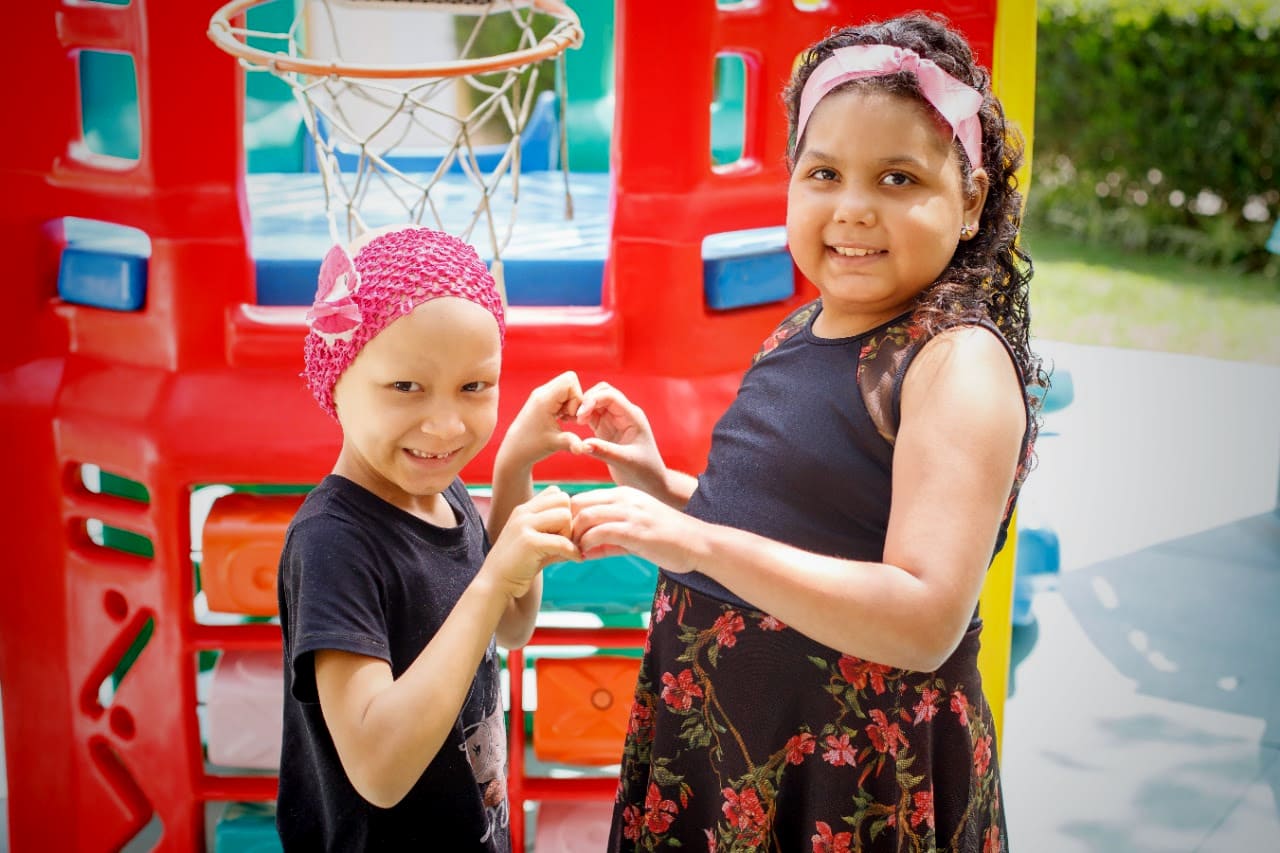 Foto colorida, em área externa, com duas crianças fazendo sinal de coração com as mãos. Uma delas está em tratamento do câncer e usa toquinha rosa na cabeça.