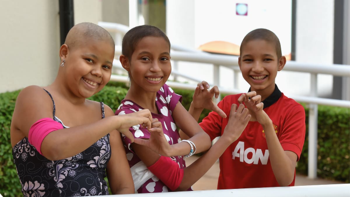 Foto com três crianças em tratamento de câncer, hóspedes da Casa Ronald McDonald. Elas sorriem, fazem coraçãozinho com as mãos, superando desafios do câncer infantojuvenil.