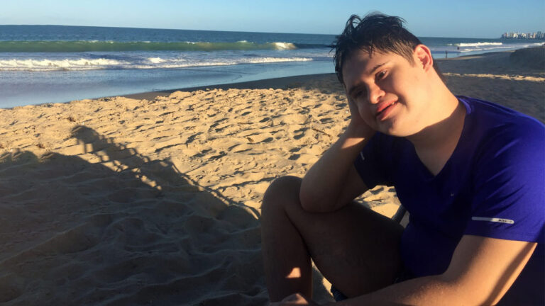 Augusto, jovem adulto sentado na praia de Boa Viagem, no Recife, com camiseta azul e sorrindo. Ilustra o lado B do autismo e síndrome de down.