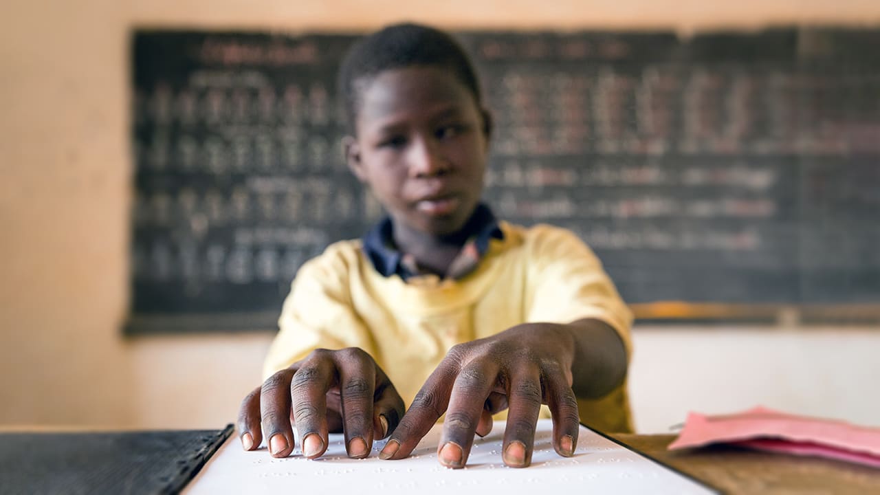 Jovem estudante negro fazendo leitura em braille dentro da sala de aula. Ao fundo há uma lousa. Dia Mundial do Braille 2023.
