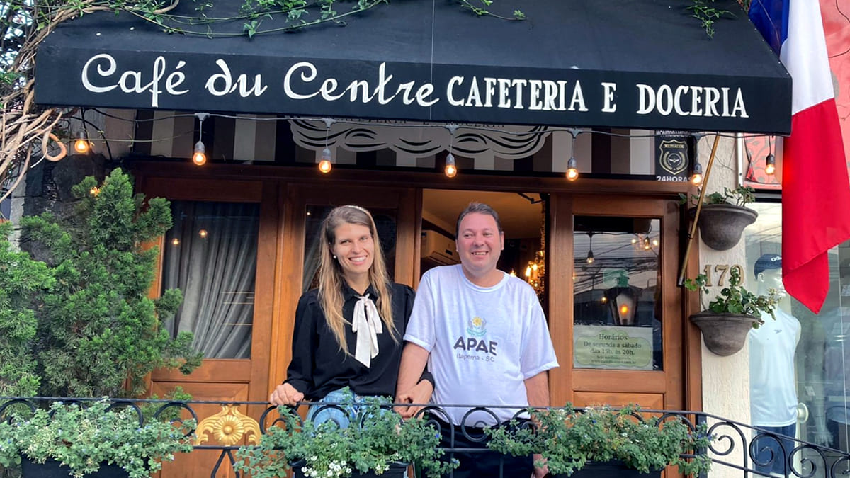 Uma das fundadoras do Café Du Centre, em foto ao lado de membro da Apae, na frente da matriz da rede de cafeterias. Os dois sorriem pela parceria Apae e cafeteria.