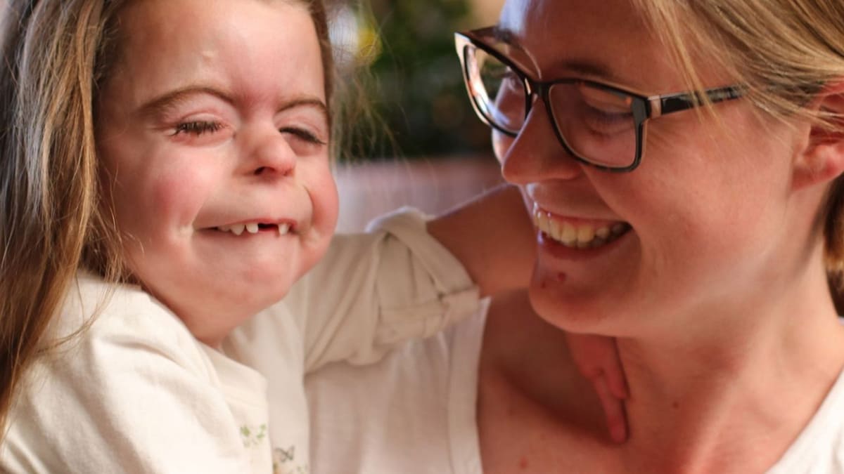 Foto de criança do grupo de doenças raras, com síndrome de Cornélia de Lange (SCdL). Ela está no colo da mãe, que usa óculos de grau. As duas são brancas, têm cabelos loiros e estão sorrindo.