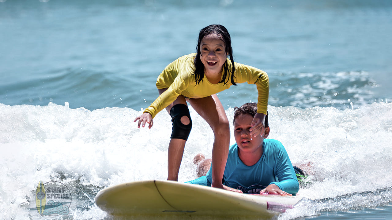 Foto de jovem com deficiência praticando o surf adaptado no Guarujá. A garota está em pé na prancha com outro jovem segurando a prancha do projeto Surf Especial.