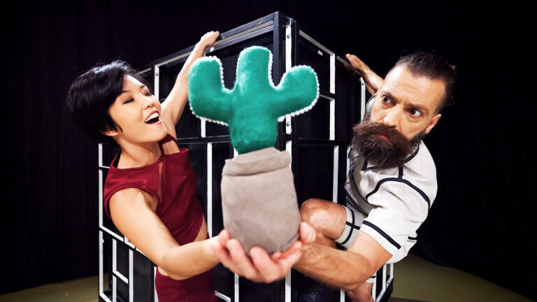 Débora Ishikawa e Alessandro Coelho durante encenação do espetáculo “Sob o Mesmo Teto”. Ambos seguram vaso com um cactos cenográfico.
