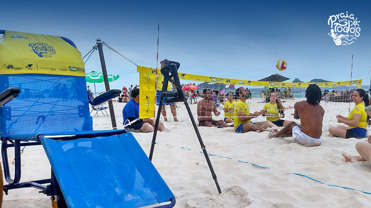 Pessoas com deficiência no desafio de vôlei sentado na areia da praia na Barra da Tijuca, Rio de Janeiro. Sobreposição da logo do projeto Praia Para Todos, no canto direito superior.