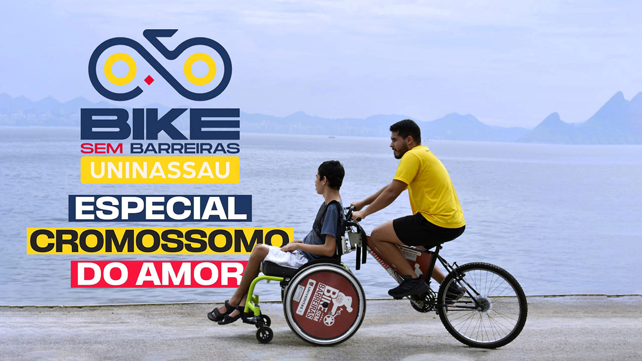 Pessoa com deficiência sentada em cadeira de rodas, adaptada na frente da bicicleta, que é conduzida por guia do projeto. Logo do Bike Sem Barreiras e do Cromossomo do Amor.