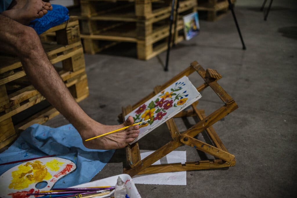 Foto de artista com deficiência pintando quadro com o pé na feira Rio Artes Manuais 2023 - Faço Arte, Faço Parte.