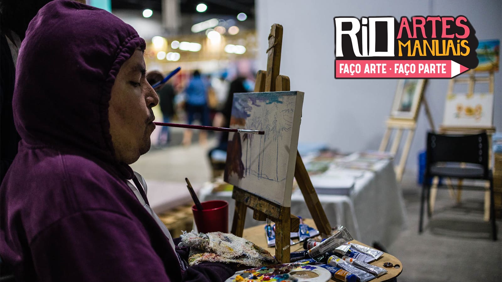 Artista com deficiência pintando quadro com a boca. Sobreposição da logo Rio Artes Manuais 2023 - Faço Arte, Faço Parte.
