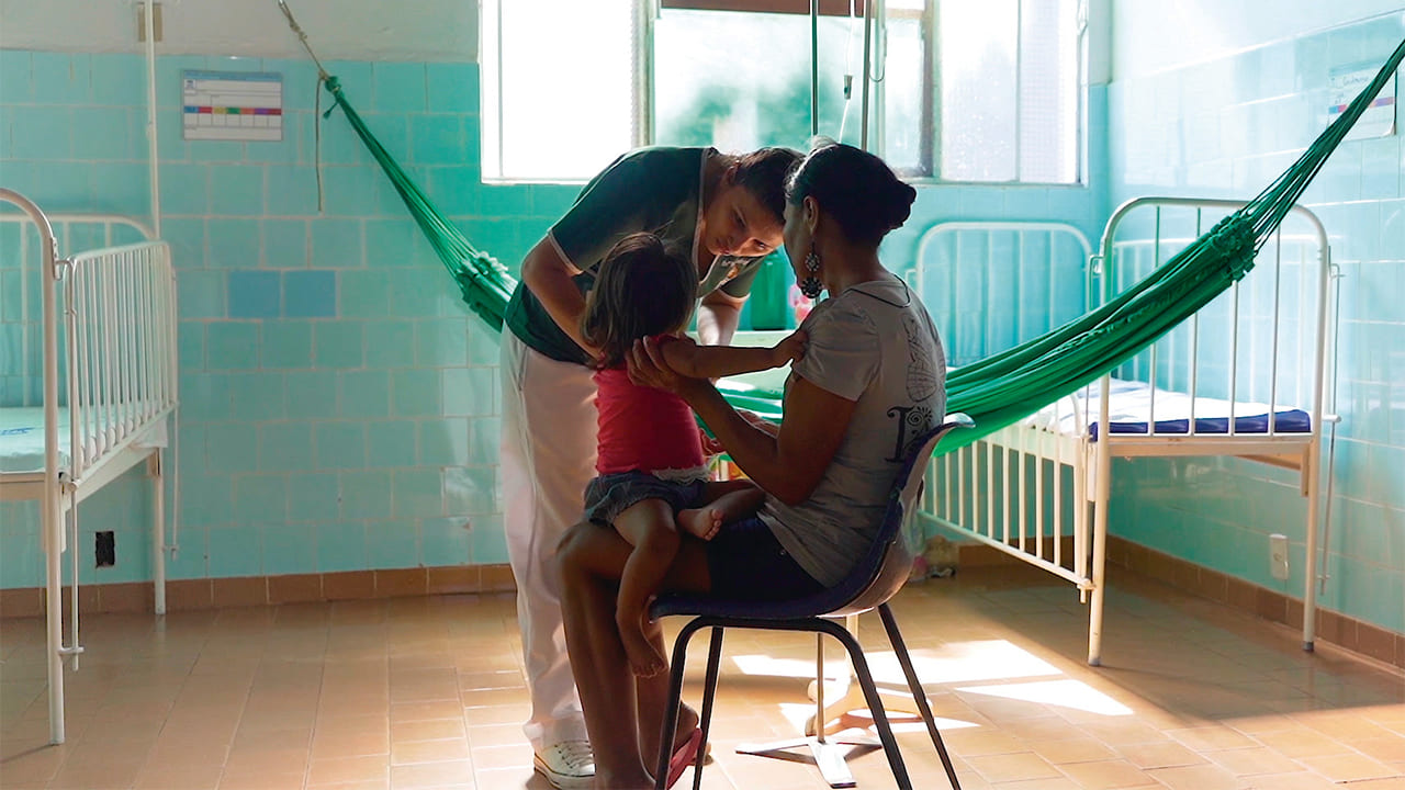 Imagem de atendimento a pacientes indígenas na enfermaria do Hospital Bom Pastor. Mulher sentada com criança no colo interagindo com enfermeira. No local há duas camas, uma rede e janela.